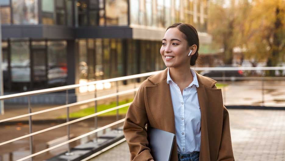 Image of young brunette asian woman wearing earbuds holding laptop computer while walking through city street